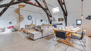 A spacious loft with exposed wooden beams, a loft bed, a dining area with a table set for six, and a kitchen in the background. - 4 Bed in Thorpeness (oc-cho) (Thorpeness)