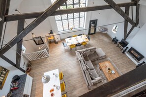 A spacious loft with exposed wooden beams, a dining area with yellow chairs, a grey sofa, and a fireplace. - 4 Bed in Thorpeness (oc-cho) (Thorpeness)