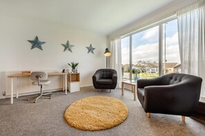 Ein modernes Wohnzimmer mit Schreibtisch, zwei Sesseln, einem runden Teppich und großen Fenstern mit Vorhängen.
