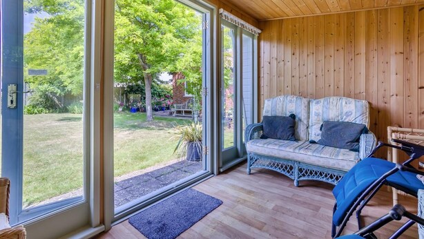 Une pièce avec un plafond en bois, un canapé en osier agrémenté de coussins, un fauteuil roulant et de grandes fenêtres donnant sur un jardin et une maison en briques.