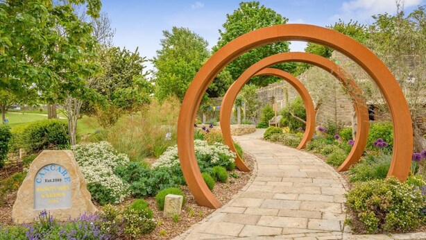 Ein gepflasterter Weg führt durch einen Garten mit einem Metallbogen und einem Schild, das einen Garten der Stille kennzeichnet.