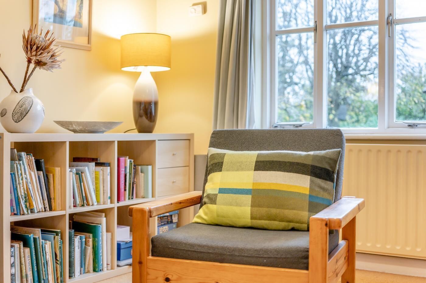 A cozy reading chair with a checkered pillow, a bookshelf filled with books, a lamp, and a window with a view of trees.
