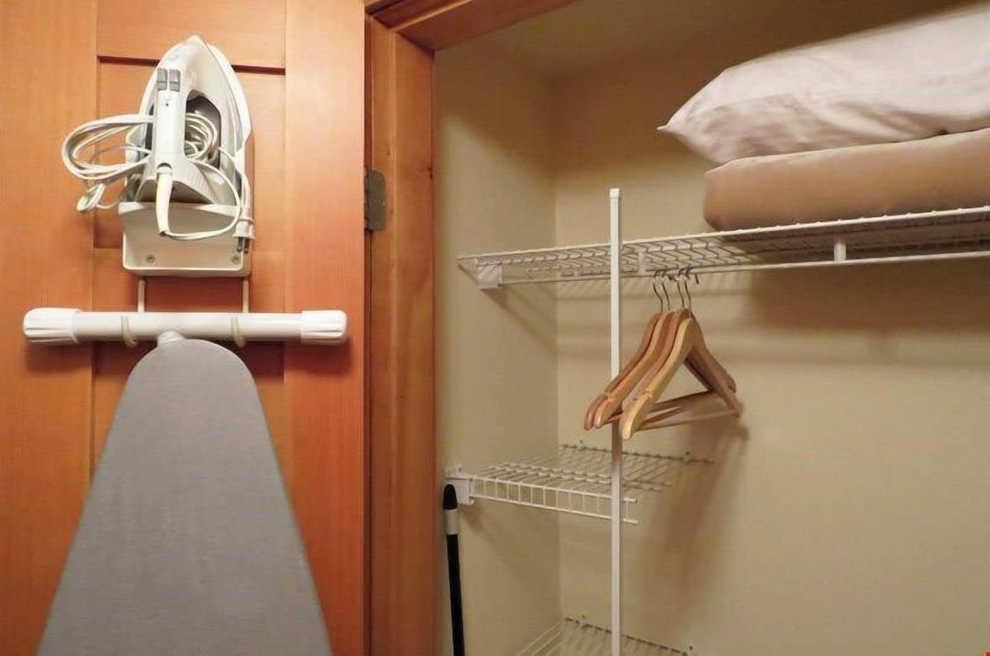 Een kleine slaapkamer met een houten deur, een wit strijkijzer, een metalen plank met een opgevouwen matras, en een hangrek.