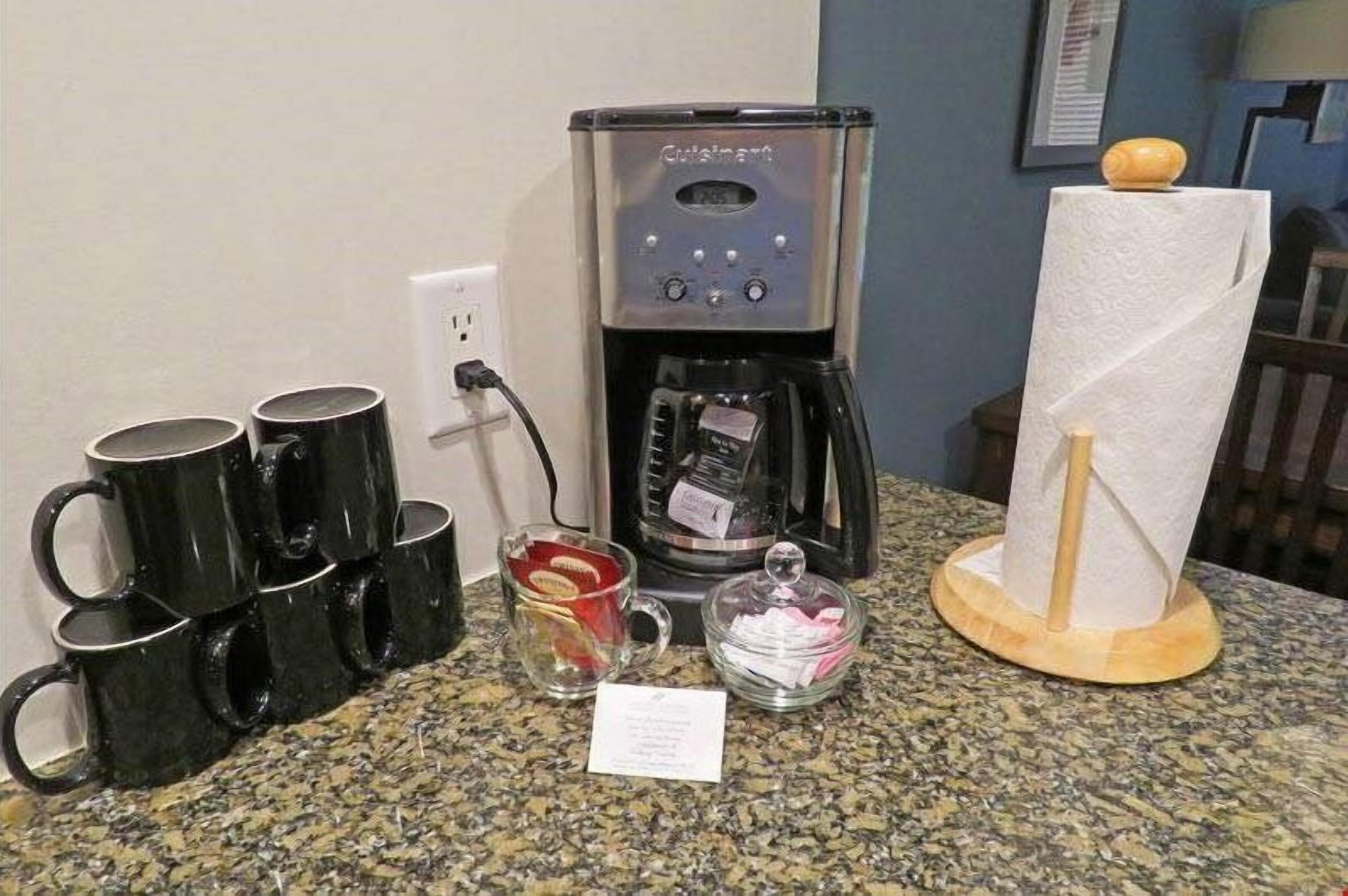 Eine Kaffeemaschine, schwarze Tassen, ein Glas mit roter Hülle, eine Tissue und eine kleine Karte auf einer Granitabdeckung.