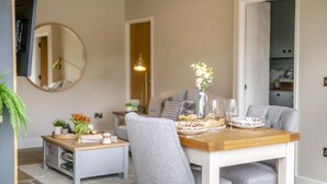 A dining area with a wooden table, grey upholstered chairs, and a vase of flowers. There is a television mounted on the wall and a mirror above the table. - 1 Bed in Askrigg (oc-c33109) (Askrigg)