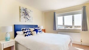 Een slaapkamer met een bed, nachtkastje, lamp, raam met gordijnen en een schilderij aan de muur.