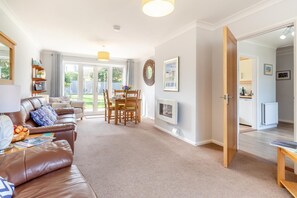 A spacious living room with a leather sofa, dining table, and chairs, a fireplace, and a view of the backyard through large windows. - 2 Bed in Reydon (oc-lowrd) (Reydon)