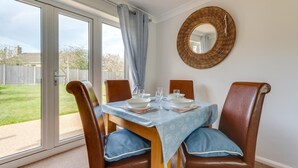 A dining area with a table set for four, a wicker mirror on the wall, and a view of a backyard through large sliding glass doors. - 2 Bed in Reydon (oc-lowrd) (Reydon)