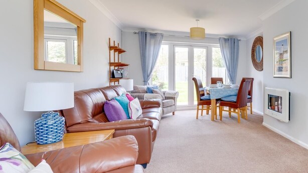 A living room with a brown leather sofa, a dining table with chairs, a lamp, and a large window with curtains. - 2 Bed in Reydon (oc-lowrd) (Reydon)