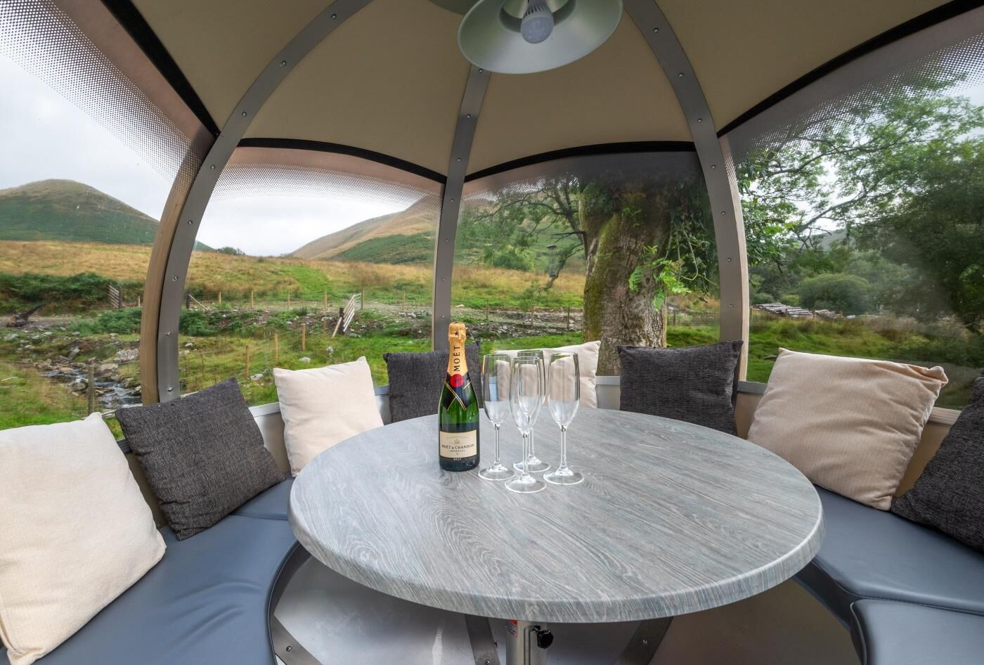 A round table with a bottle of champagne and glasses, surrounded by cushioned seating with pillows.