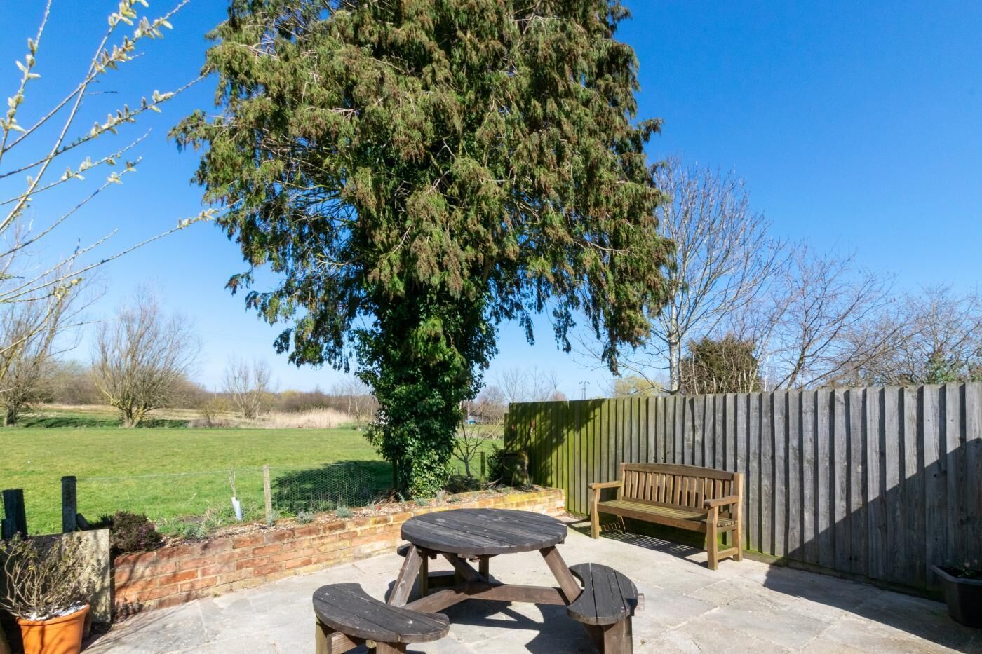 A patio with a wooden table and benches, a wooden fence, and a large tree in the background.