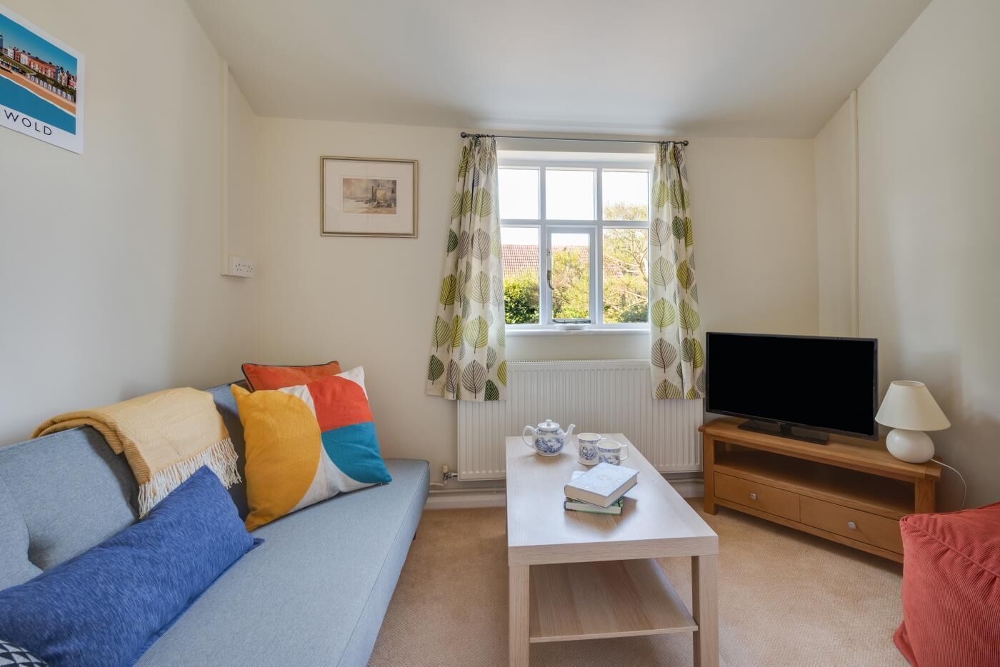 A living room with a sofa, a television, a table with a book and teapot, and a window with curtains.