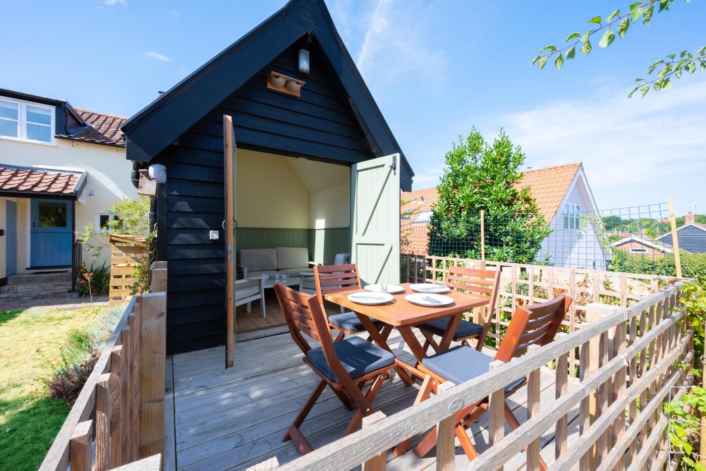 A wooden deck with a table and chairs, a small black building with a door, and a fenced garden area.
