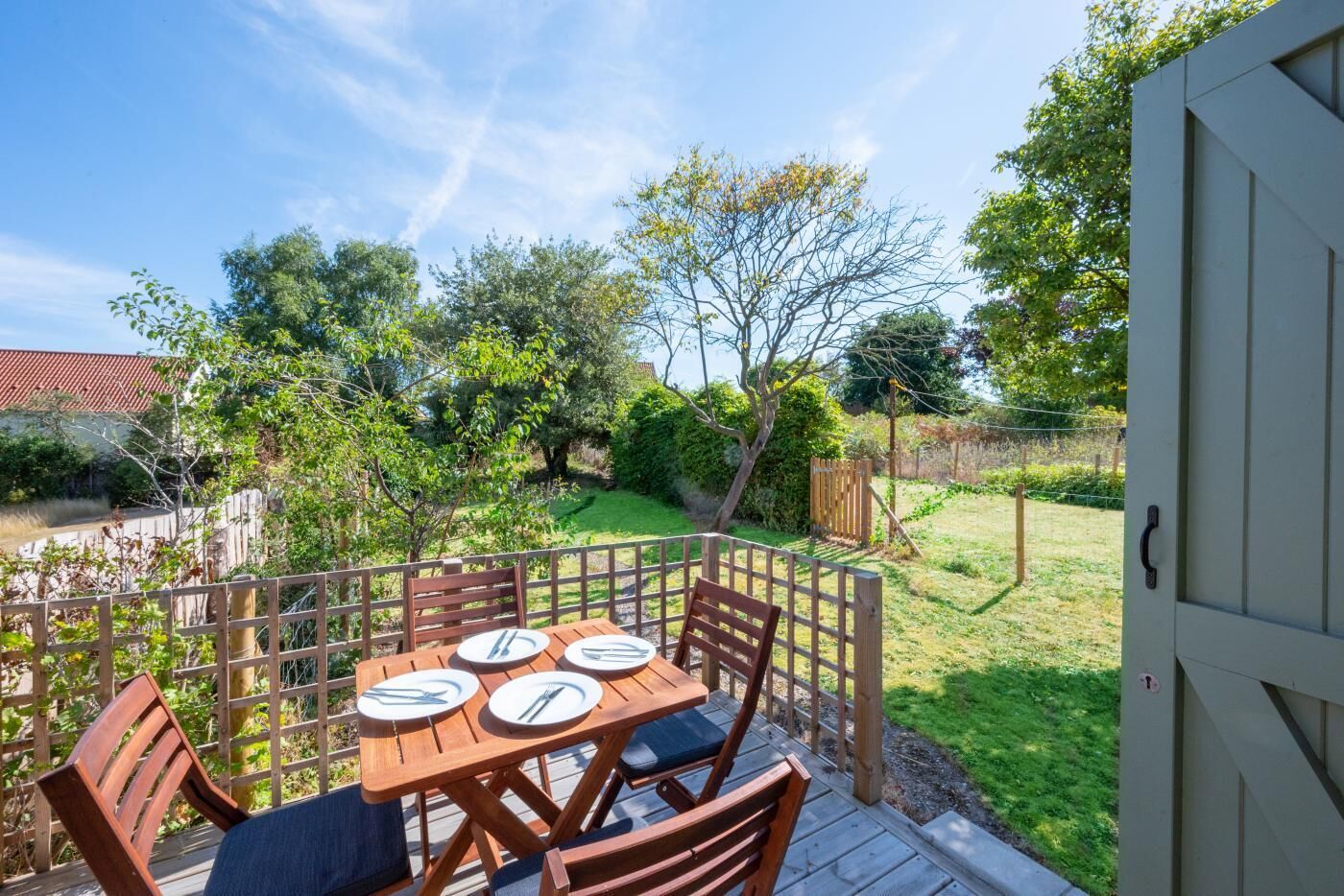 A wooden deck with a table set for four, surrounded by a wooden fence, and a garden with various trees and shrubs.