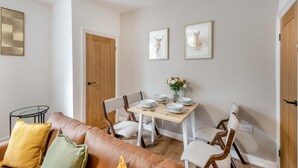 A dining area with a wooden table set for four, surrounded by white chairs. There is a brown leather couch with cushions in the foreground. - 3 Bed in Skipton (oc-g33088) (Skipton)