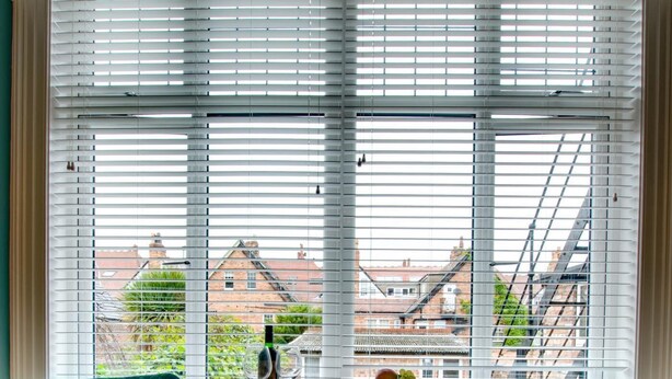 Una habitación con dos sillas color turquesa, una mesa con un frutero y una botella, y vistas a edificios a través de ventanas amplias con persianas.