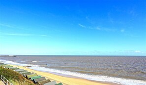 Ein Strand mit einer Reihe von Strandhäusern mit grünen Dächern entlang der Küste, ein Pier in der Ferne und ein klarer, blauer Himmel.