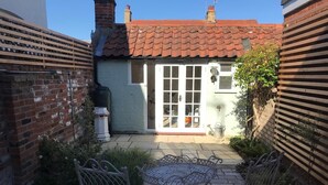 A patio with two metal chairs and a table, a brick wall, a white building with a tiled roof, and a wooden fence. - 1 Bed in Woodbridge (oc-jsm) (Woodbridge)