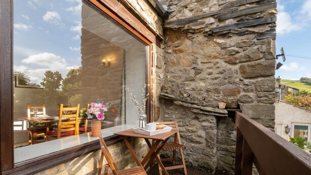 A wooden deck with a stone wall, a table with chairs, and a window overlooking a garden.