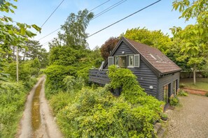 A small house with a sloped roof, surrounded by dense greenery and a narrow path leading to it. - 2 Bed in Woodbridge (oc-owls) (Ufford)