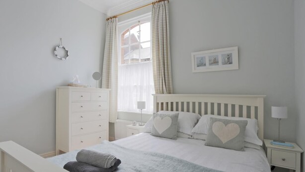 A bedroom with a bed, a dresser, a nightstand, a lamp, a window with curtains, and a wall-mounted picture. - 2 Bed in Southwold (oc-che) (Southwold)