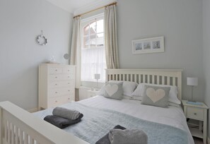 Une chambre à coucher comprenant un lit, une commode, une table de chevet, une lampe, une fenêtre avec des rideaux et un tableau accroché au mur.
