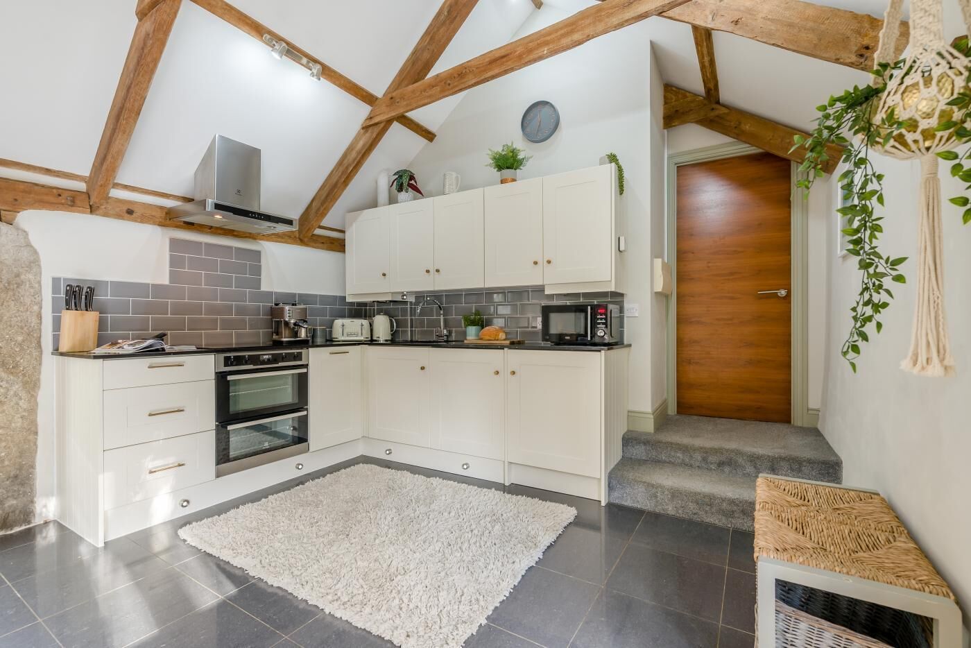 A modern kitchen with white cabinets, a built-in oven, and a microwave. There is a white rug on the floor, and wooden beams on the ceiling.