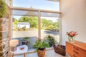 Ein Zimmer mit großem Fenster, Steinwand, kleinem Tisch mit Lampe, Korbwäsche und Holzschrank.