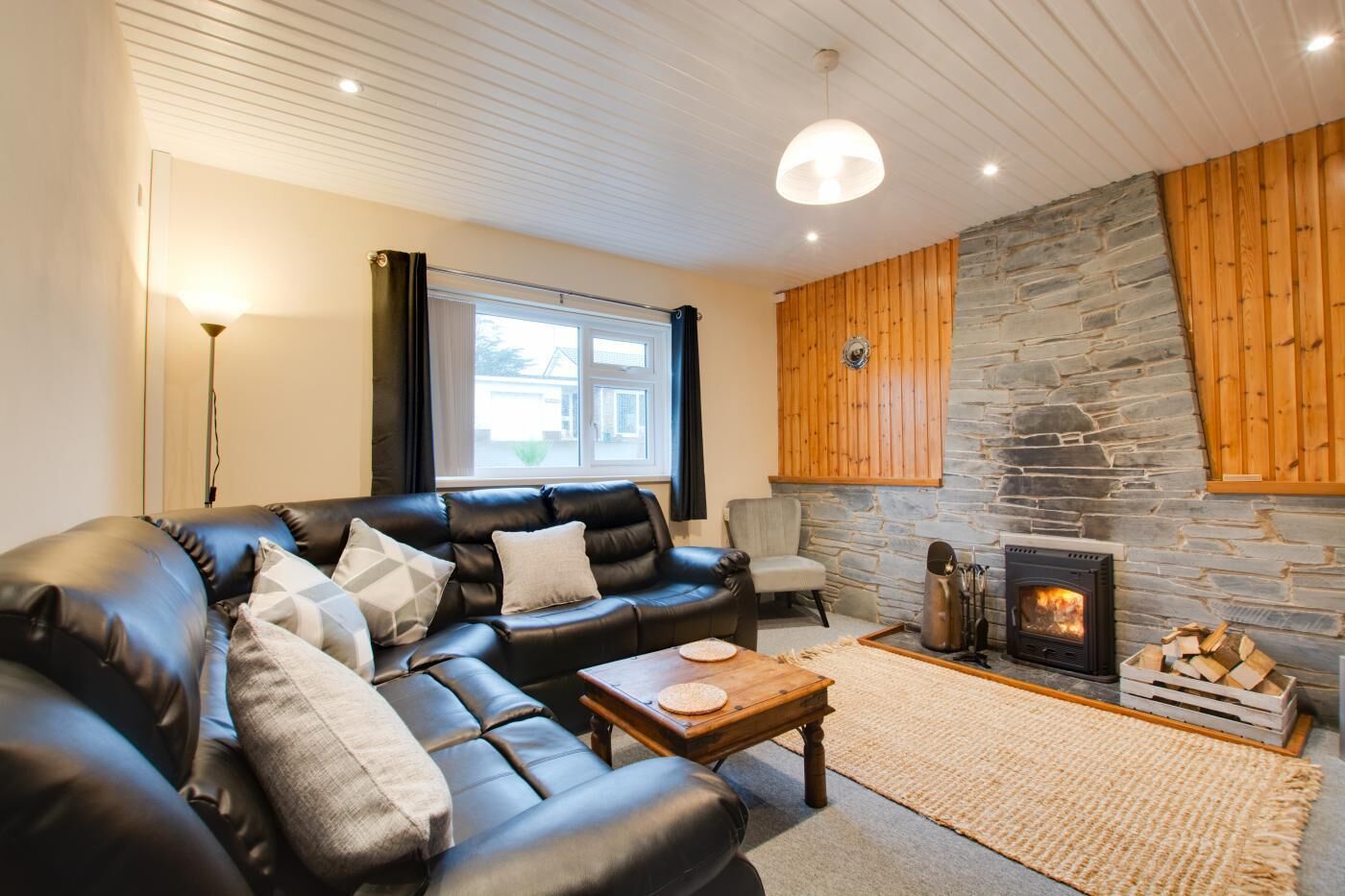A cozy living room with a black leather sofa, a wooden coffee table, a stone fireplace, and a large window with curtains.