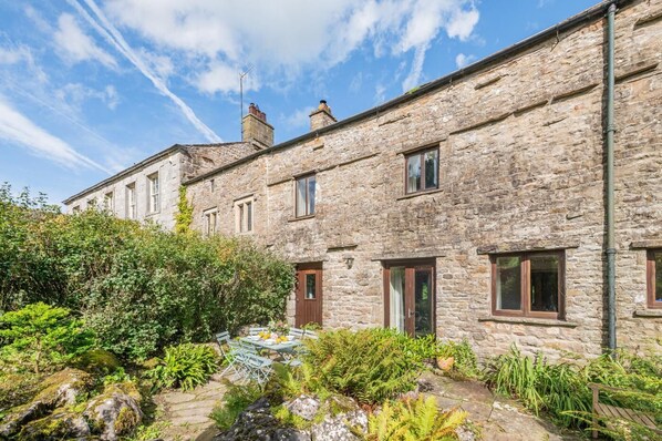 A stone building with multiple windows and doors, surrounded by lush greenery and a stone pathway. - 4 Bed in Hawes (oc-g33122) (Hawes)