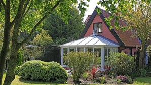 A conservatory with a glass roof, surrounded by well-maintained garden and a red-roofed house. - 1 Bed in Frostenden (oc-phc) (Frostenden)