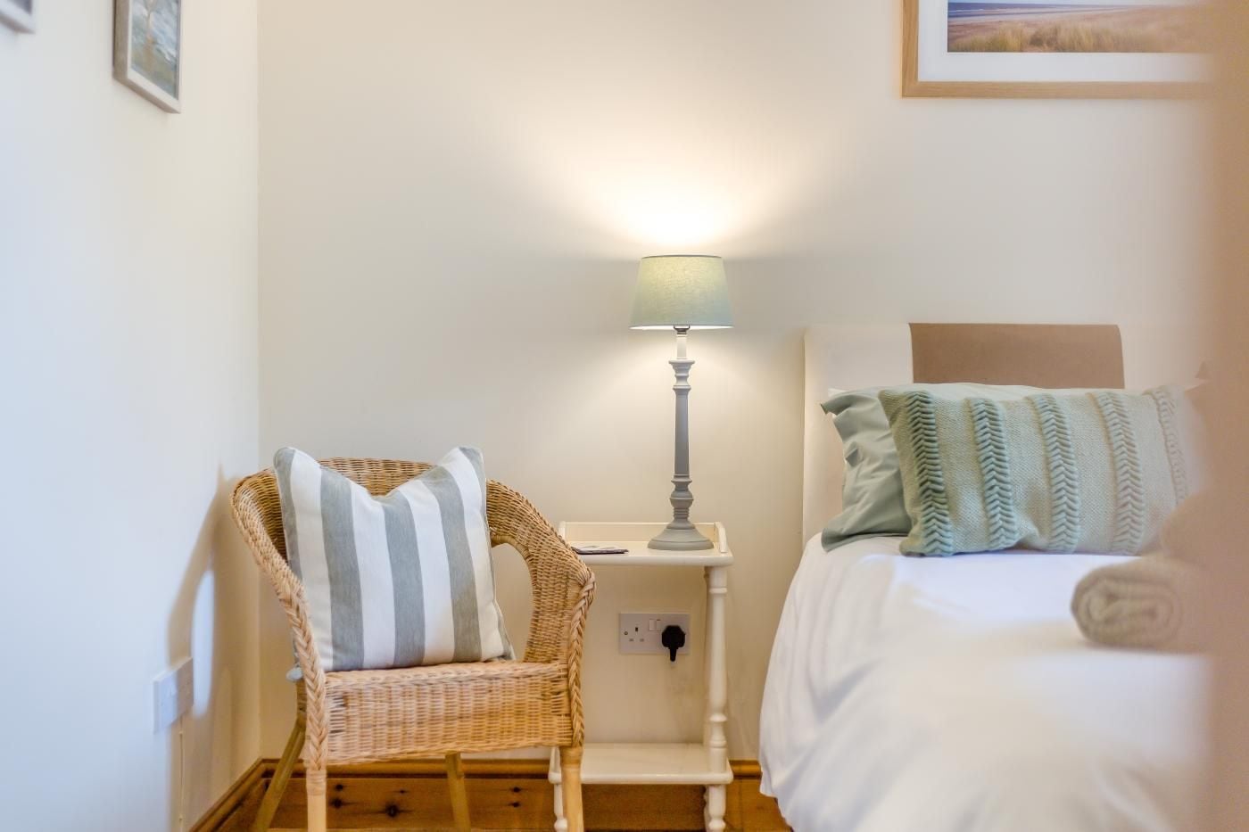 A bedroom with a bed, a bedside lamp, a wicker chair with a striped pillow, and a wall-mounted power outlet.