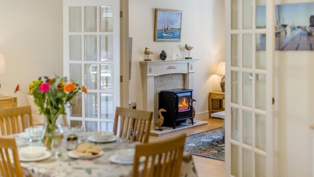A dining area with a table set for four, a fireplace with a wood-burning stove, and a view of the sea through a window. - 2 Bed in Blakeney (oc-1558) (Blakeney)