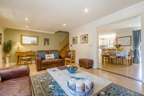 Una sala de estar con un sofá de cuero marrón, una mesa de centro con objetos decorativos, una escalera y un comedor con mobiliario de madera.