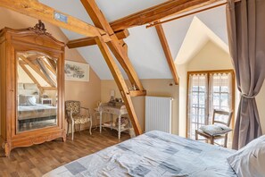 Ein Schlafzimmer mit schrägen Wänden, Holzbalken, einem großen Spiegel, einem Bett, einem Sessel, einem kleinen Tisch und einem Fenster mit Vorhängen.