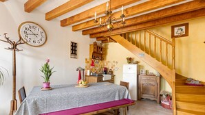 Un espace repas comprenant une table, des chaises, un banc, une horloge, un lustre, un escalier et, au fond, une cuisine.