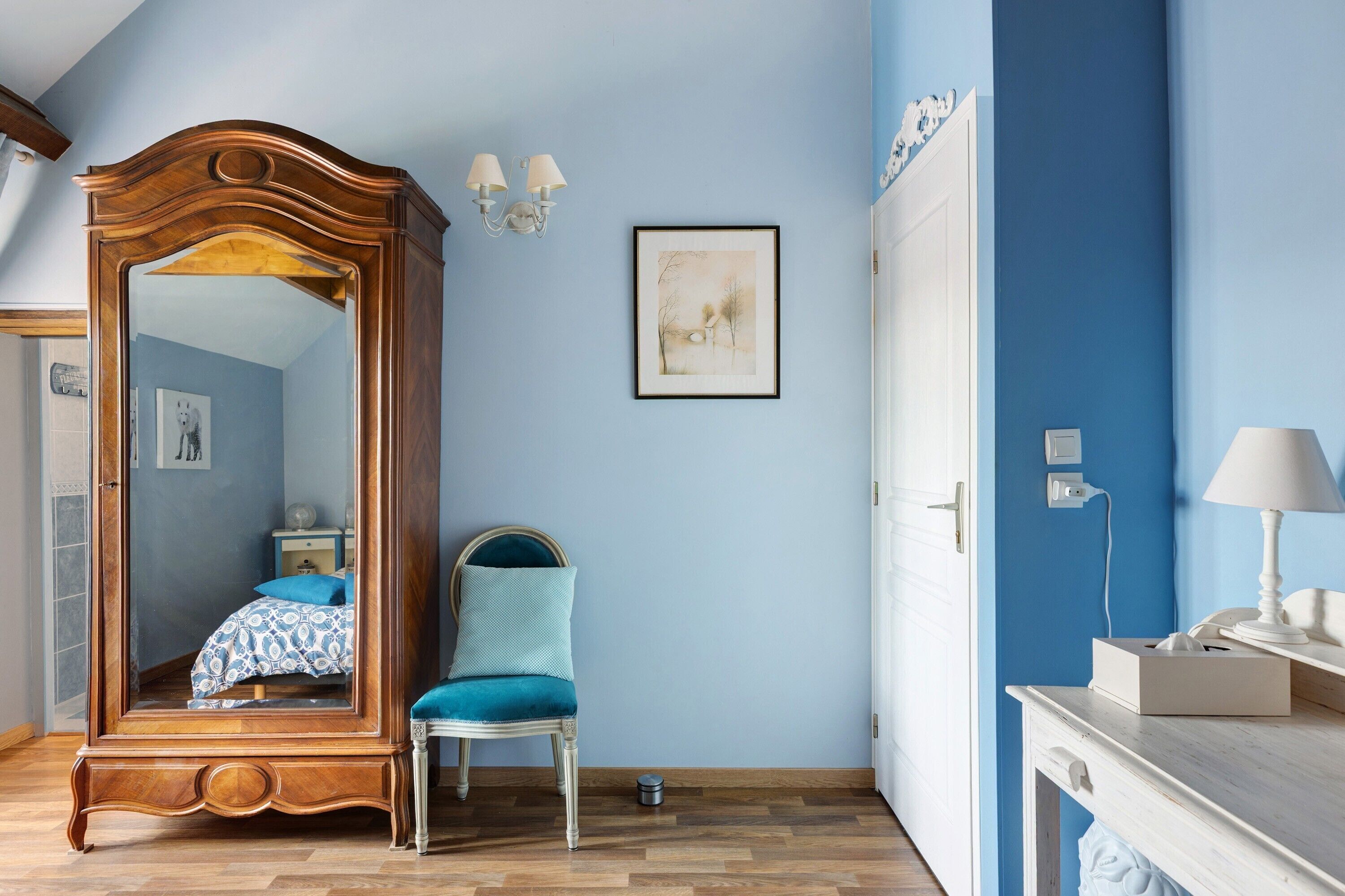 Une chambre avec un grand miroir, un lit recouvert d’une couvre-lit à motifs bleus et blancs, un fauteuil recouvert d’un tissu turquoise, une table de chevet blanche avec une lampe, et un tableau encadré accroché au mur.