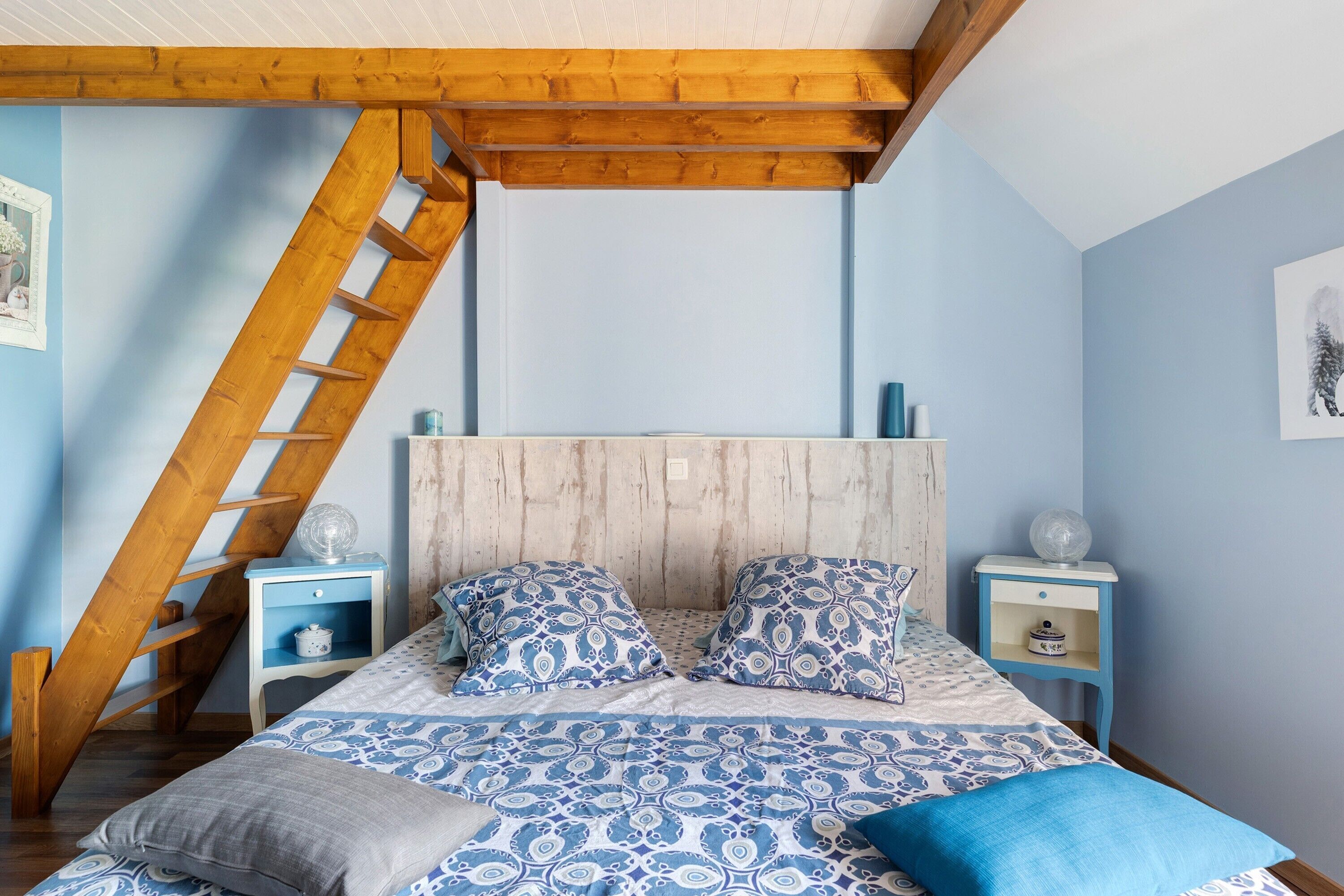 A bedroom with a sloped ceiling, wooden beams, a bed with patterned bedding, two bedside tables, and a ladder.