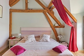 A bedroom with a wooden ceiling, a bed with white and pink bedding, two bedside tables with lamps, and a red curtain.