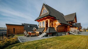 Une cabane en rondins avec un toit à forte pente, un porche à l’avant avec des marches et un petit espace extérieur aménagé pour s’asseoir.