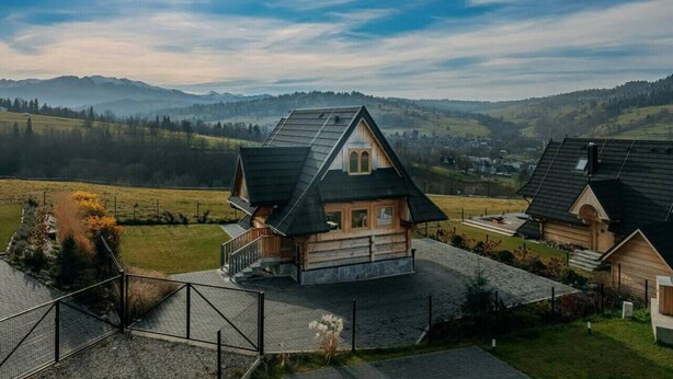 Une maison en bois, dotée d’un toit à forte pente, entourée d’une cour clôturée et offrant une vue dégagée sur les collines environnantes.