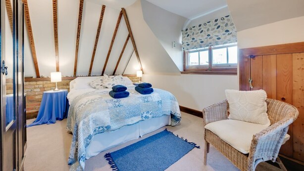 A bedroom with a sloped ceiling, a bed with blue bedding, a wicker chair, and a window with patterned curtains. - 1 Bed in Woodbridge (oc-hok) (Bawdsey)