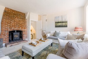 Uma sala de estar com lareira de tijolos, sofá branco, tapete estampado e um quadro na parede.