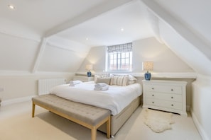 Quarto com teto inclinado, cama com roupa de cama branca, um banco, cômoda e duas luminárias.