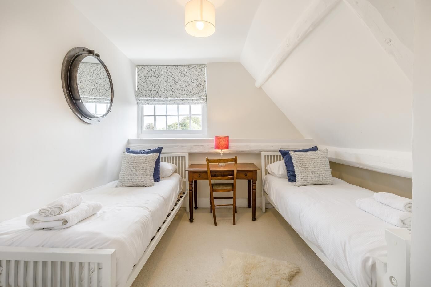 A small, well-lit room with two single beds, a desk, and a chair.