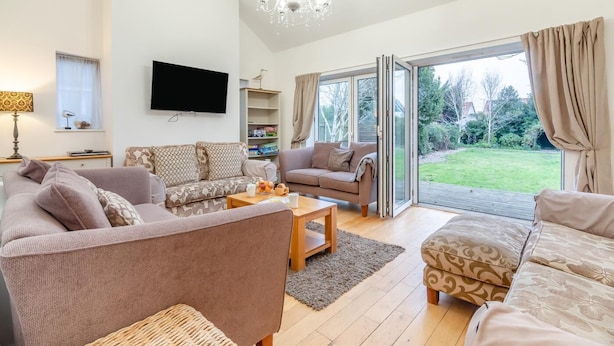 Een ruime woonkamer met een grote zithoek, een salontafel en een televisie. Er is een boekenkast en een raam met gordijnen.