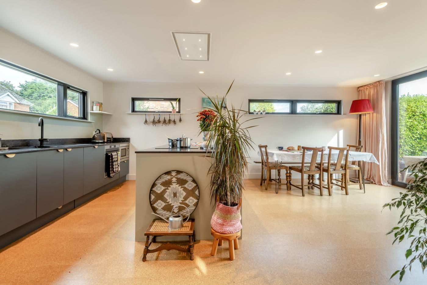 A modern kitchen with a central island, dining area with wooden chairs, and large windows providing natural light.