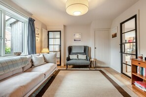 Een gezellige woonkamer met een bank, fauteuil en boekenkast, met een groot raam en een deur naar buiten.