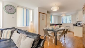 A modern living room with a black leather sofa, a dining table with chairs, and a kitchen area with white cabinets. - 3 Bed in Southwold (oc-ftw) (Southwold)