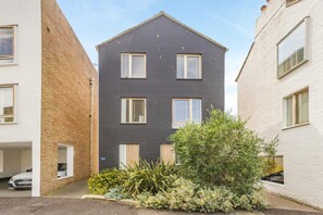 Un immeuble résidentiel moderne à deux étages, doté d’une façade sombre, de nombreuses fenêtres et d’un petit jardin agrémenté de diverses plantes.